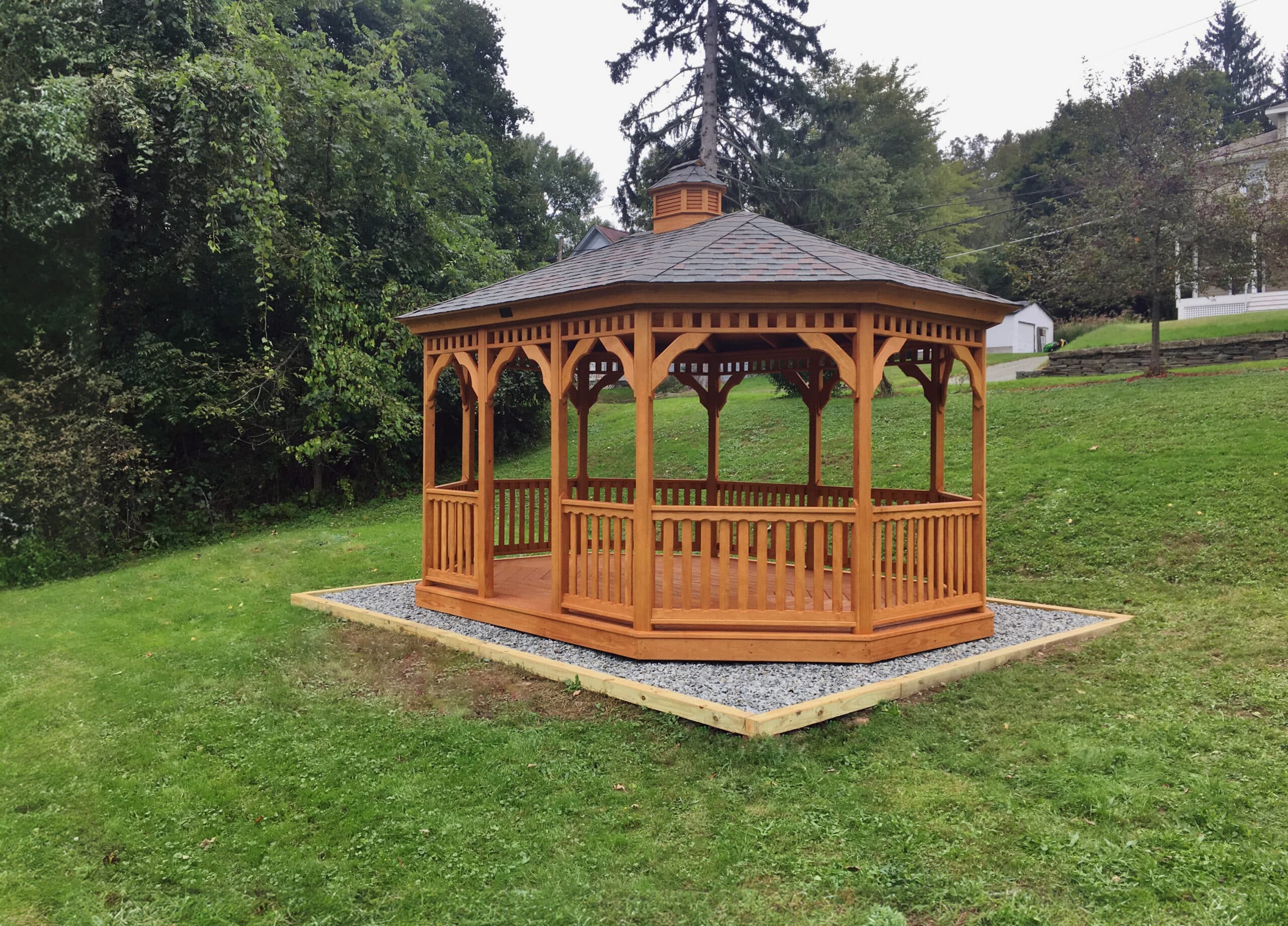Wooden oval gazebo in yard, placed on stone pad on cloudy day