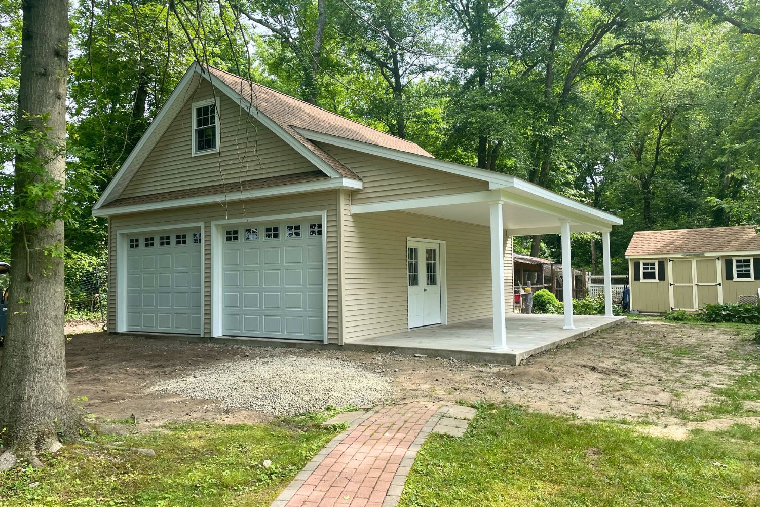 A-Frame Attic garage with brick path in woods with beige siding and white doors and trim