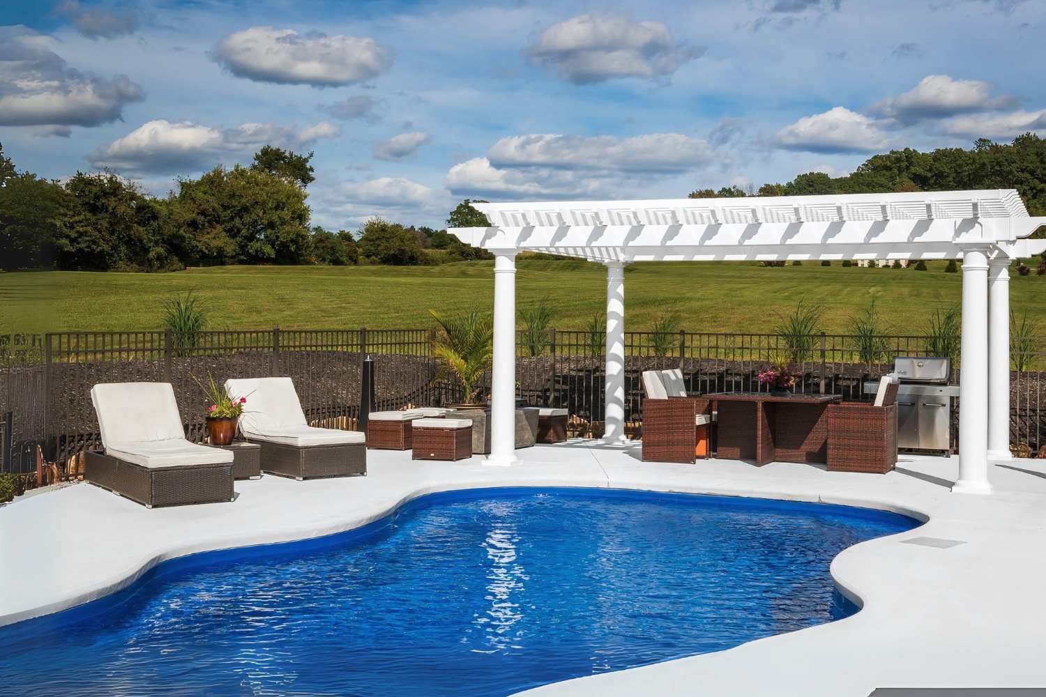 View of Cancun Pergola besides pool with seating underneath and besides pergola on a partly cloudy, bright day