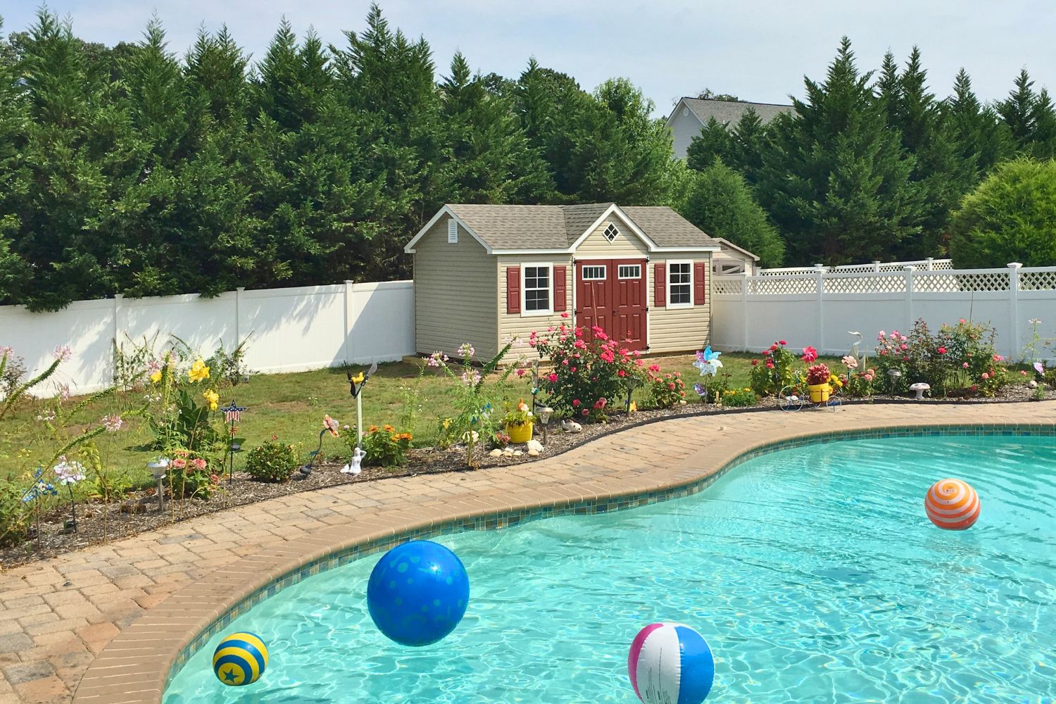 Beige cape cod shed with red doors and shutters and white trim across from pool and garden