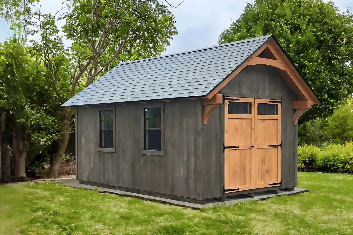 lancaster timber frame shed with gray siding and shingle roof featuring double wooden doors with transom windows. Shed on right side of image.